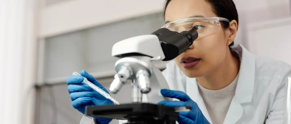 A healthcare researcher wearing protective eye goggles, gloves, and a lab coat looks into a miscroscope.